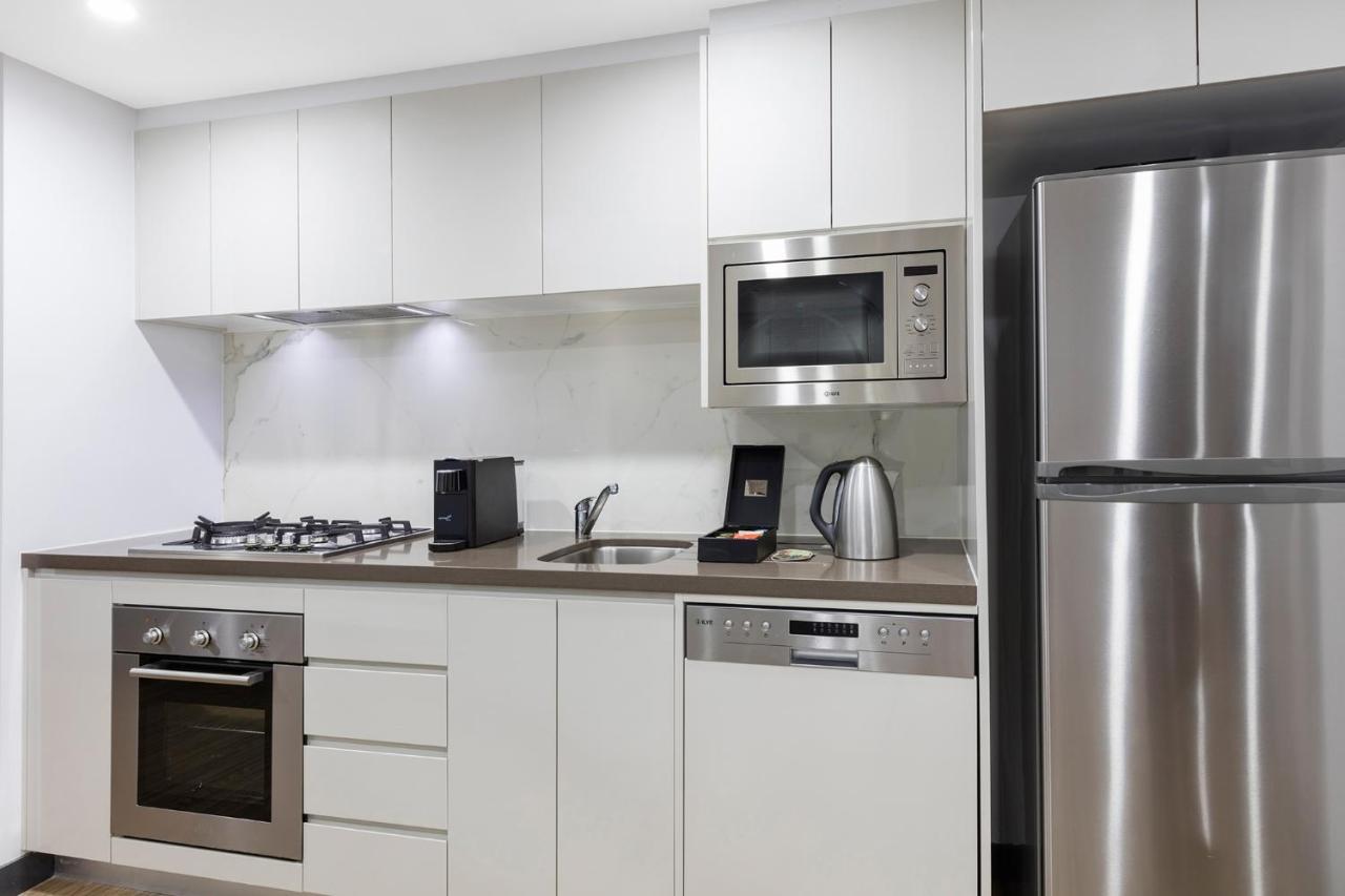 Meriton Suites Bondi Junction Sydney Exterior photo The photo shows a modern kitchen with a sleek, minimalist design. It features white cabinetry and a countertop that blends with the overall aesthetic. 

Key elements include:

- A stainless steel refrigerator on the right.
- An oven with a gas cookto